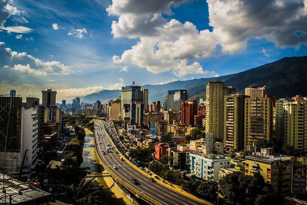 Cityscape of Caracas, Venezuela, with tall buildings and a highway running through the center. Mountains are in the background under a cloudy sky.