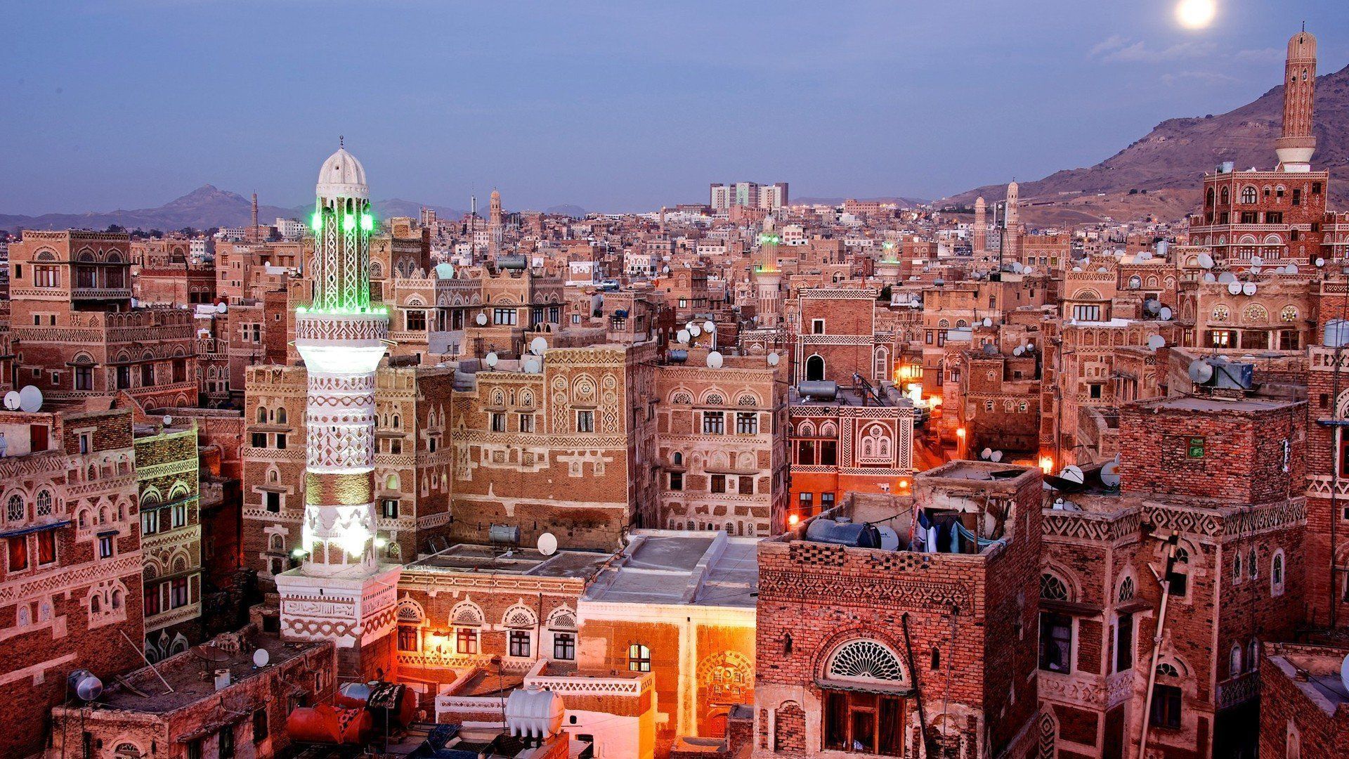 A cityscape in Yemen at dusk, featuring brown, ornate buildings, minarets, and a mountain backdrop under a dusky sky and moon.