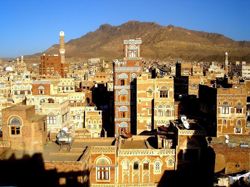 Cityscape in Sana'a, Yemen, with tall, ornate brick buildings and a mountain in the background under a clear sky.