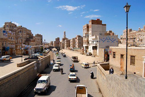 Street scene in Sana'a, Yemen, with cars and motorcycles on the road, surrounded by traditional multi-story buildings, under a bright blue sky.