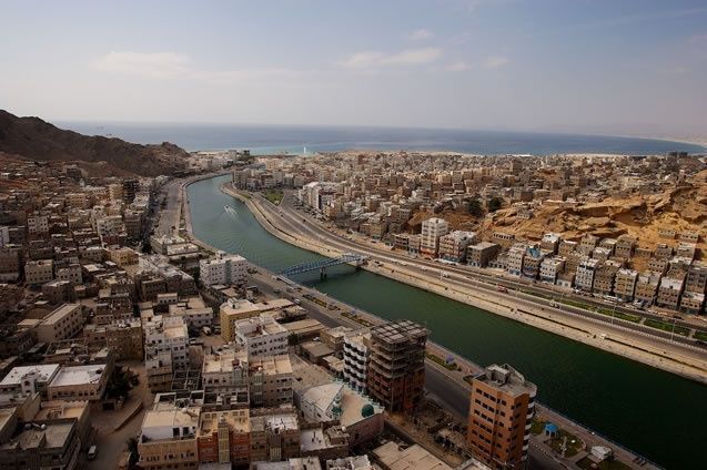 Aerial view of a coastal city with a river flowing through it, surrounded by buildings and mountains.