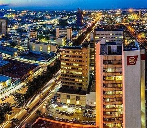 Cityscape at dusk with illuminated buildings, long exposure of car lights on road, central tower with a red logo.