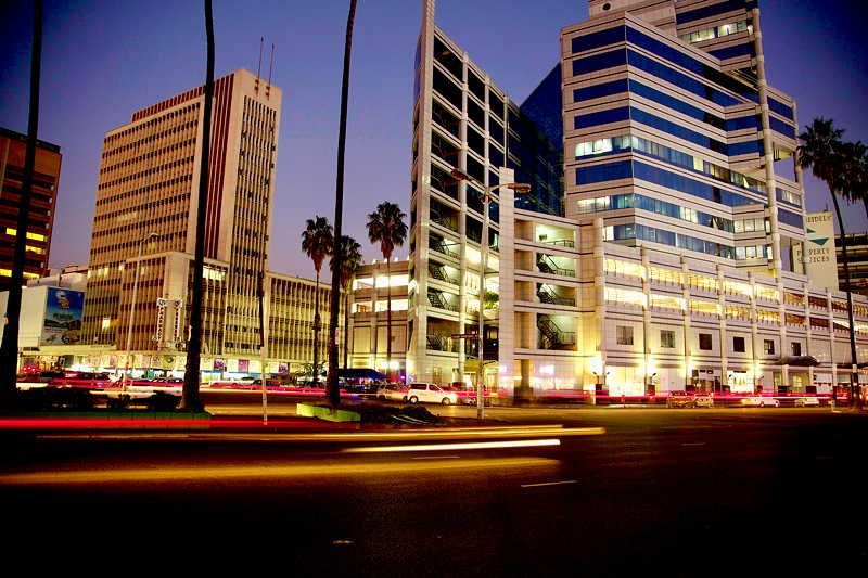 Night view of city buildings lit up with traffic streaks on a street lined with palm trees.