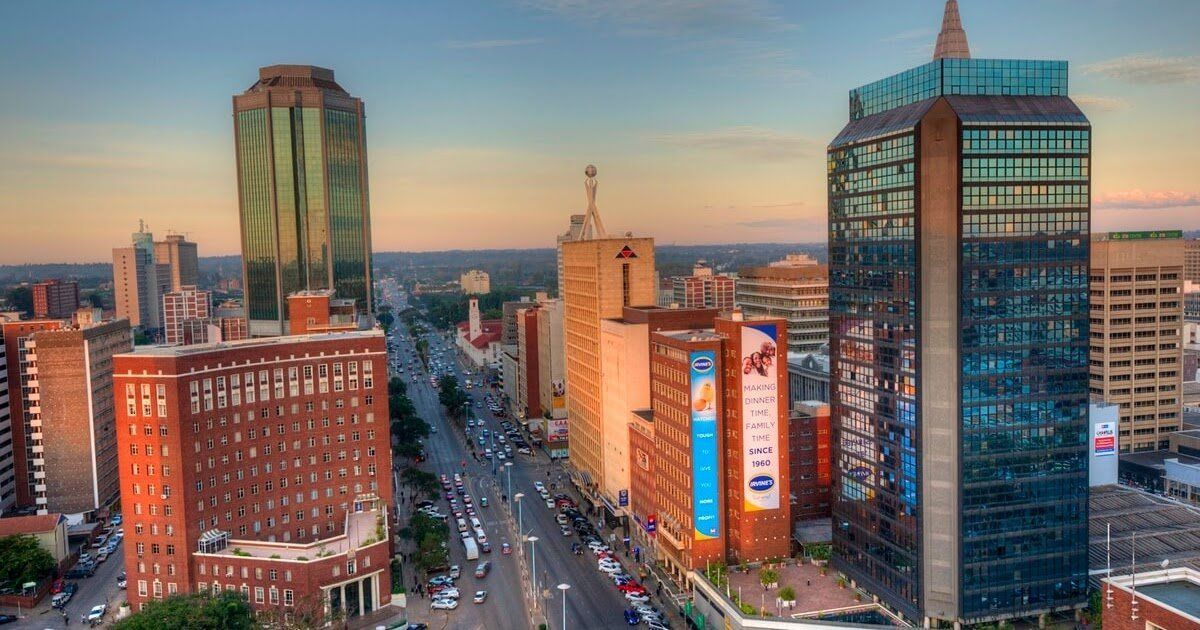 City skyline of Harare, Zimbabwe, with several tall buildings lining a wide road. The sky shows the colors of a sunset.