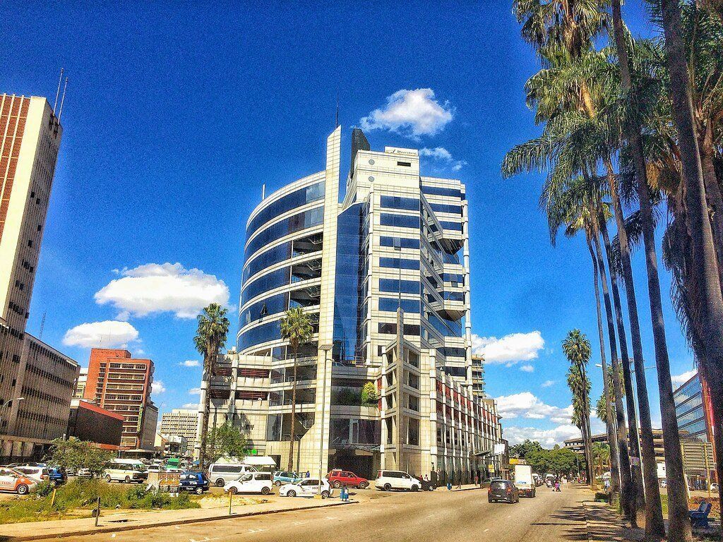 Modern skyscraper in a city, blue sky with white clouds, palm trees line the street, cars and buses on the road.