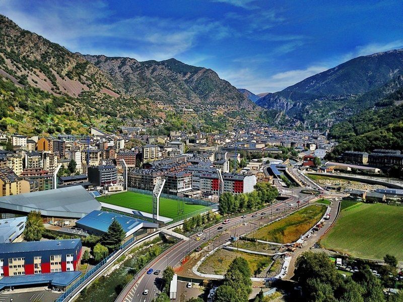 City nestled in a valley surrounded by mountains under a blue sky. Buildings and a stadium are visible, with roads winding through.