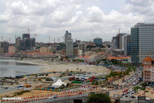 A coastal cityscape in Angola with construction cranes, high-rise buildings, a beach, and a road with traffic.