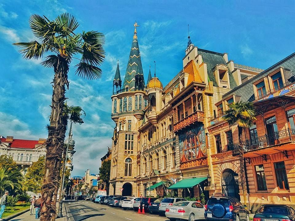 A street in Batumi, Georgia, featuring ornate buildings, palm trees, and parked cars under a blue sky.