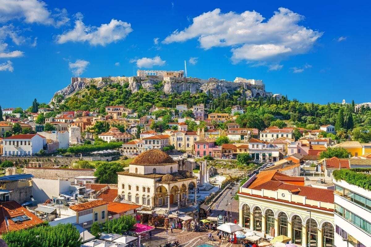 Athens, Greece cityscape with the Acropolis on a hilltop, buildings with orange roofs, and a blue sky with clouds.
