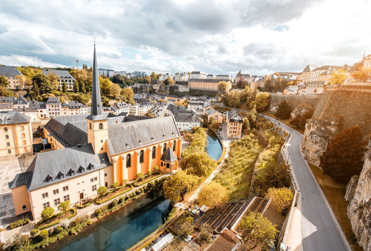 View of Luxembourg City with a church, river, and buildings nestled in a valley under a cloudy sky. A road runs along the right side.