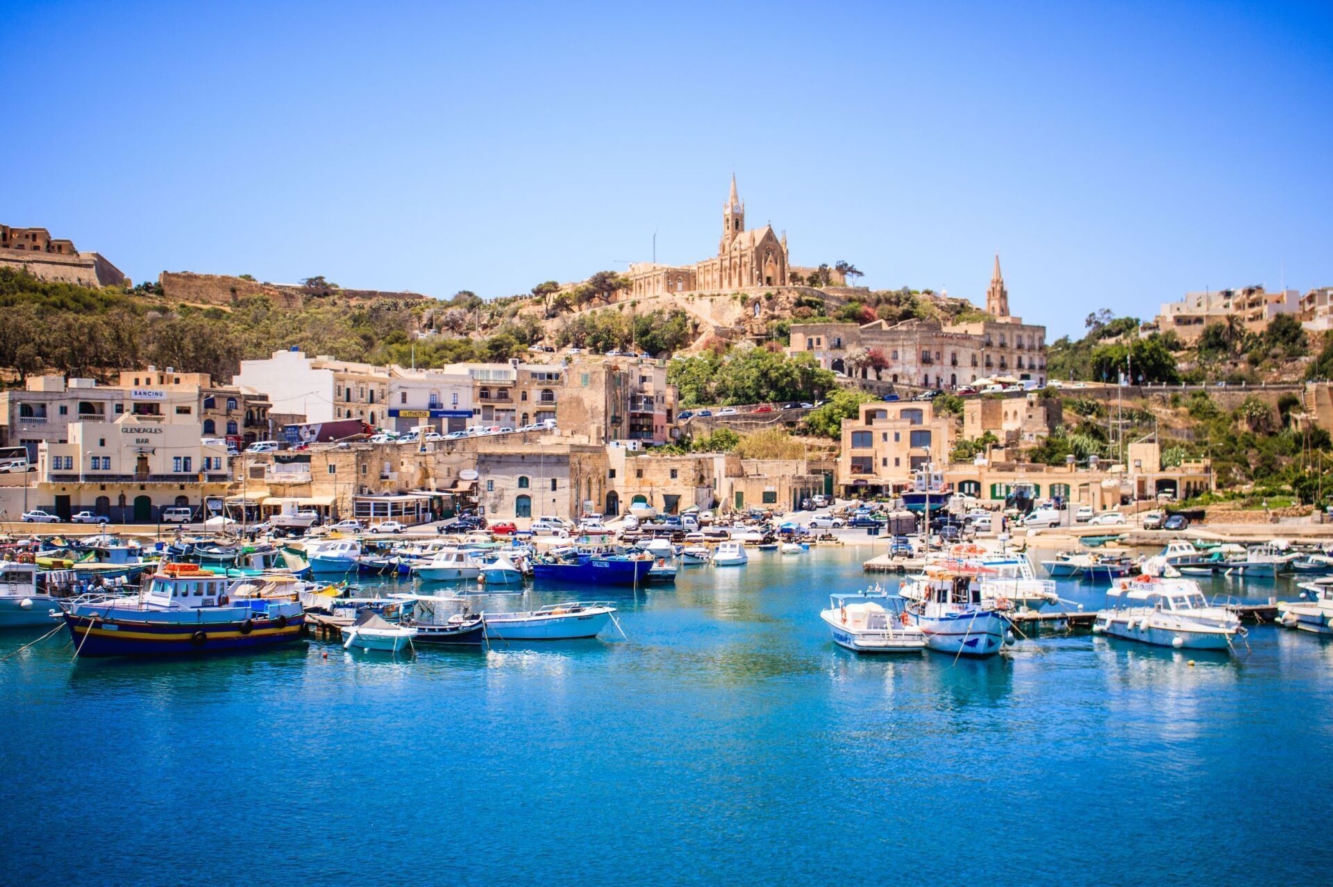 Boats docked in a harbor with buildings and a church on a hilltop under a bright blue sky.
