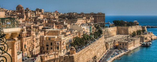 A panoramic view of Valletta, Malta. The city features beige buildings, stone fortifications, and the blue Mediterranean Sea.