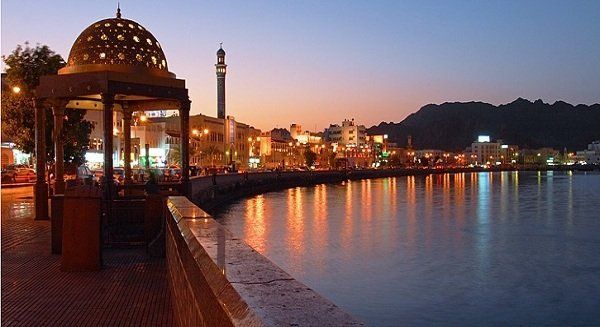 Night view of a coastal city in Oman. Buildings and mosque tower reflect in calm water at dusk.