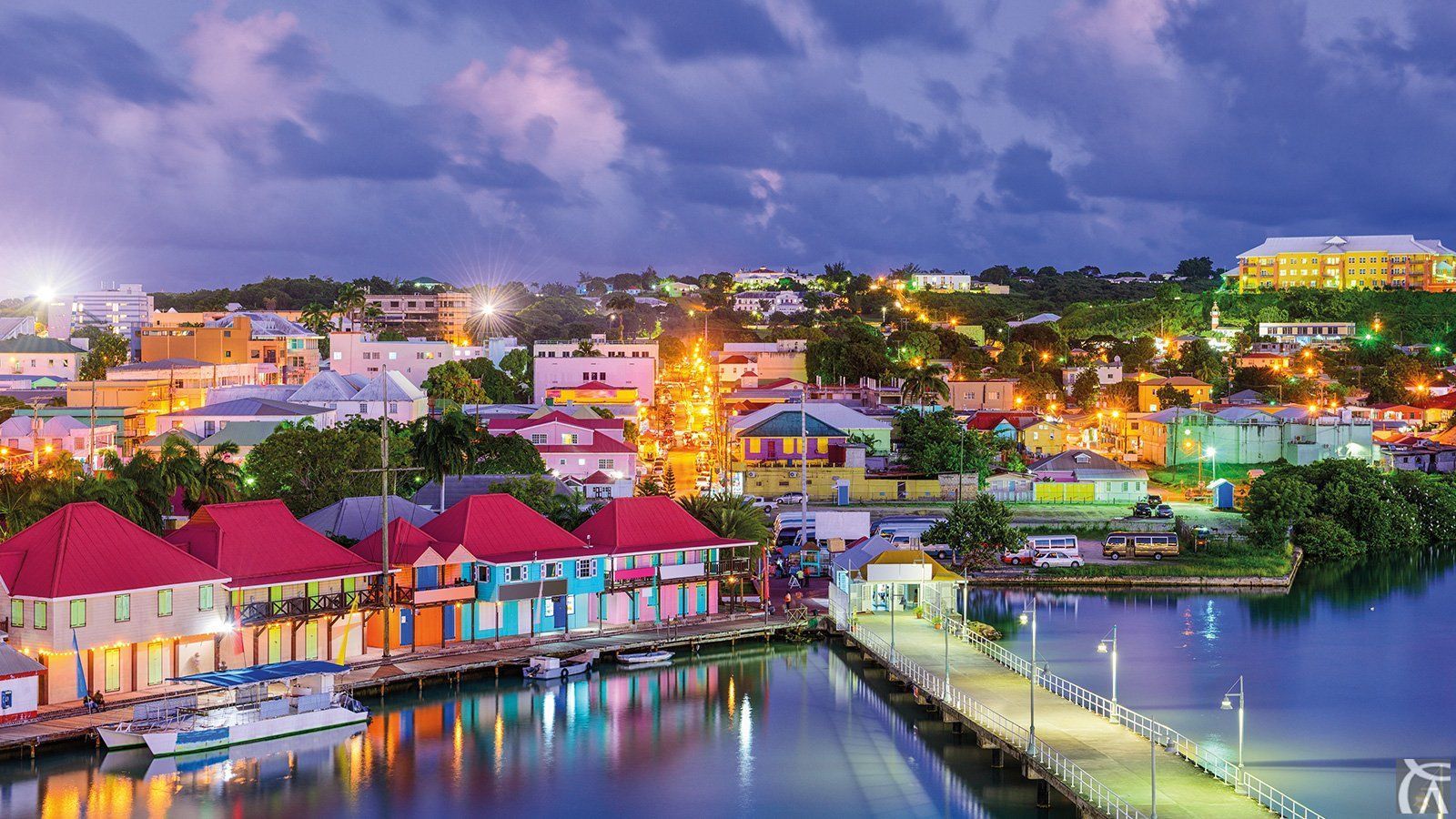 Night view of a Caribbean town with colorful buildings along a waterfront. Bright lights illuminate the buildings and a pier.