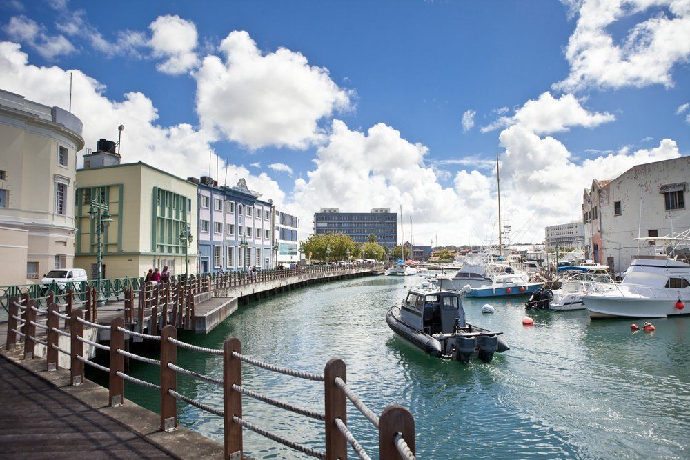 Boats on a turquoise canal in Bridgetown, Barbados, with colorful buildings and a sunny, cloudy sky.