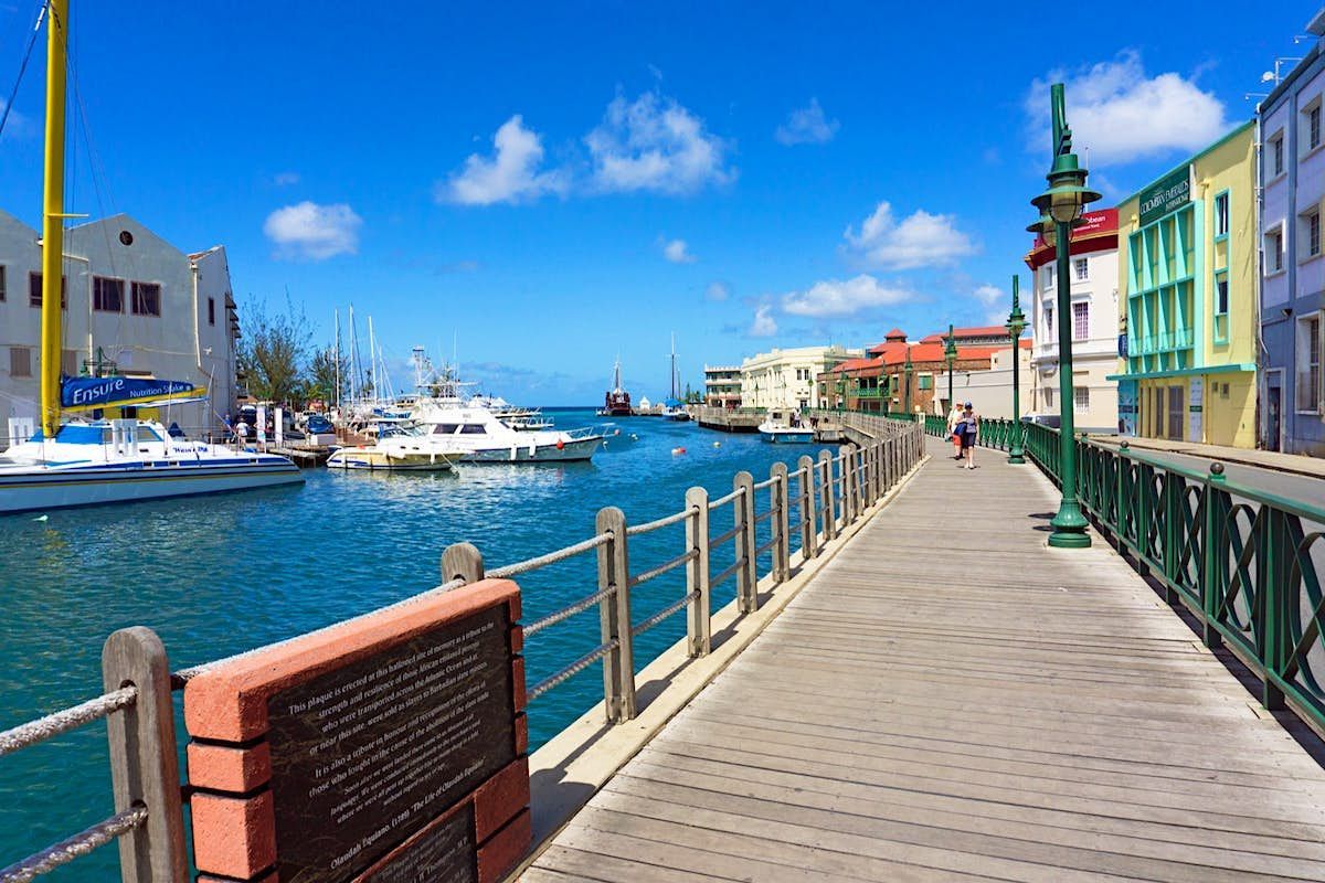 Waterfront scene in Barbados with boats docked, a pedestrian walkway, and colorful buildings under a blue sky.