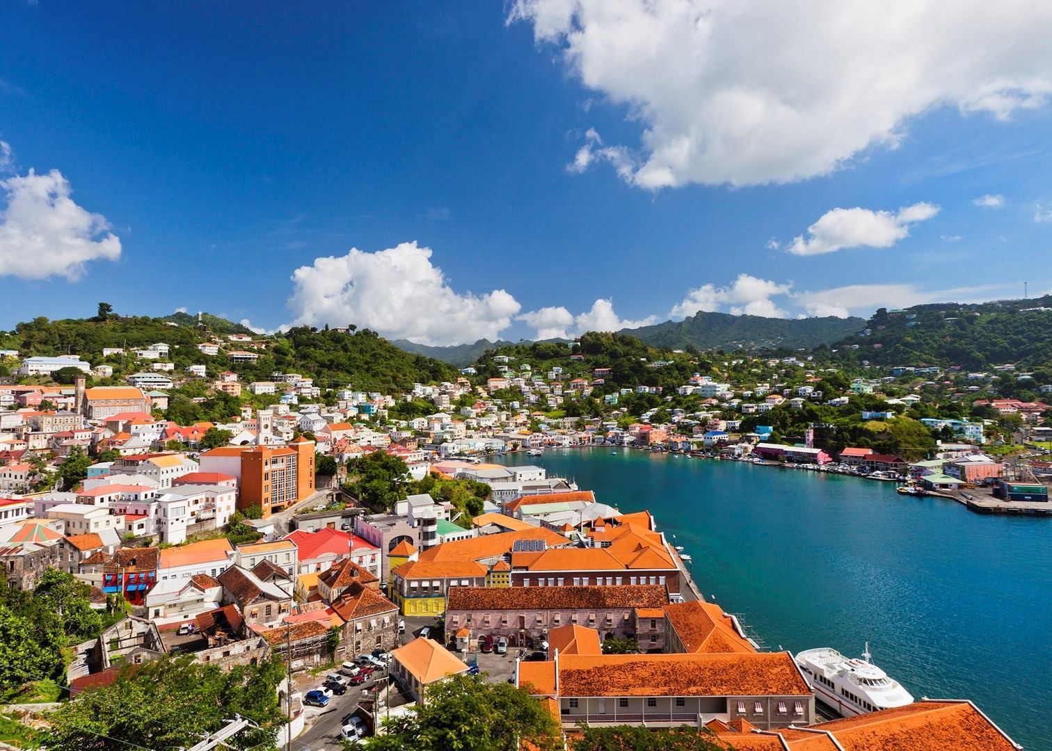 Coastal town of St. George's, Grenada, featuring buildings with orange roofs, turquoise water, and a blue sky with clouds.