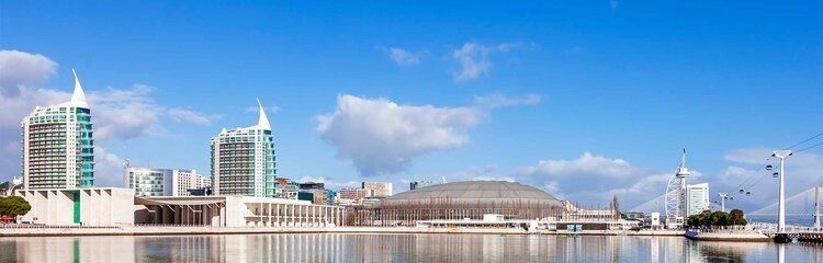 A waterfront view of buildings with spired tops against a clear blue sky. The buildings reflect in the water.