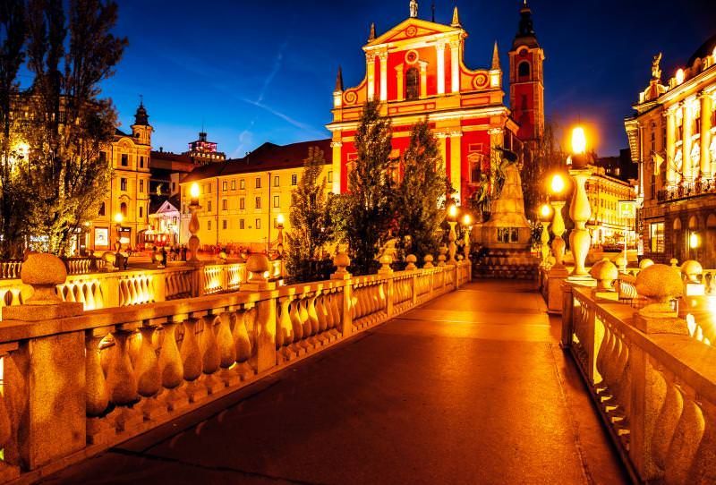 Nighttime view of Ljubljana, Slovenia. A red church and buildings line a bridge illuminated by warm streetlights and reflecting in the water.