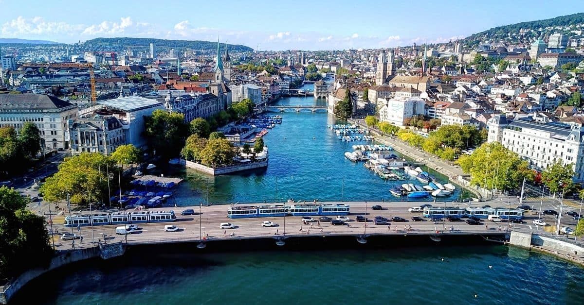 Aerial view of Zurich, Switzerland, with the Limmat River cutting through the city, bridges, boats, and buildings visible under a blue sky.