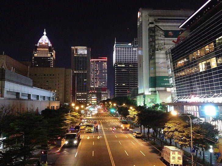 Nighttime cityscape of Taipei with illuminated skyscrapers lining a wide road, cars and streetlights visible.