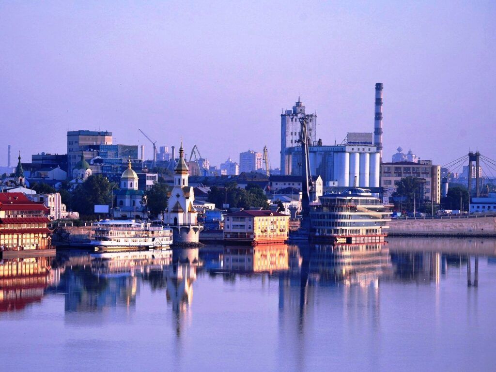 City skyline reflected in a calm river; includes buildings, boats, and a smokestack, bathed in soft morning light.