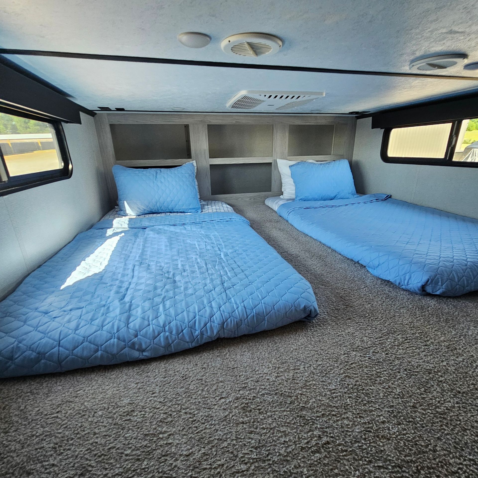 Two twin beds with light blue bedding in a room with gray carpet. There are windows on either side.