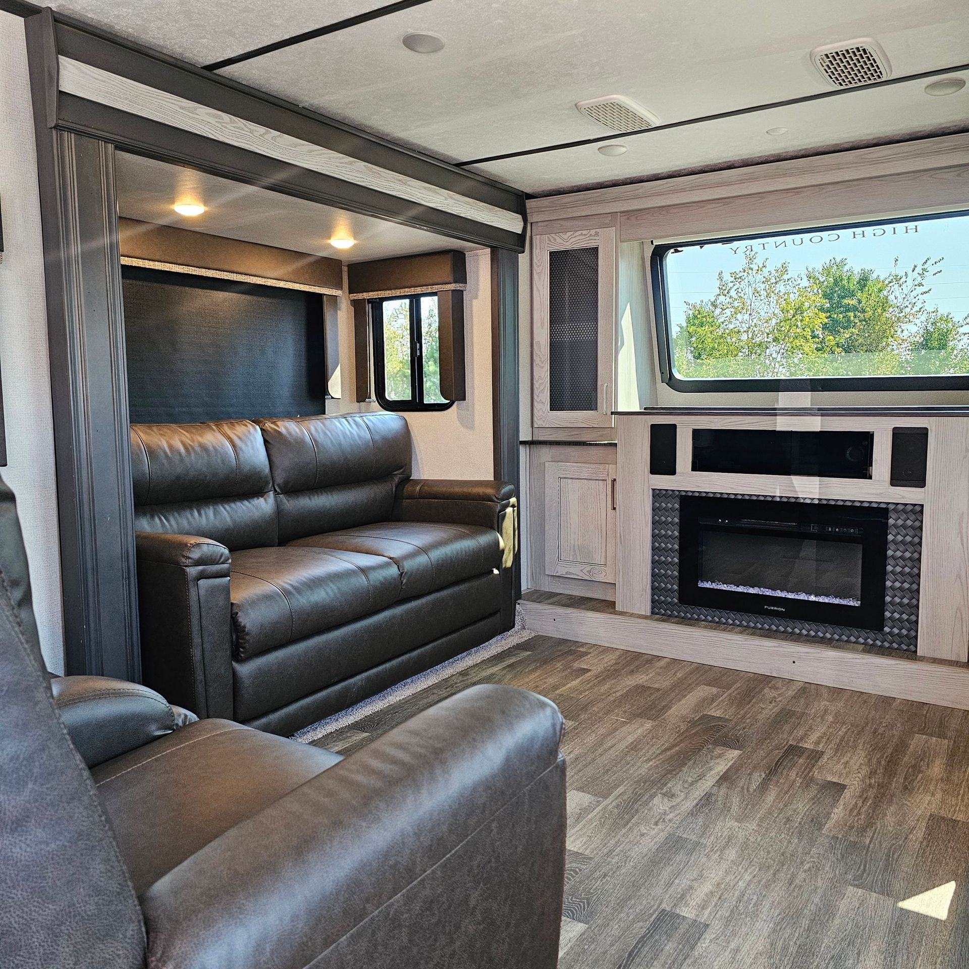 Interior of an RV featuring a leather sofa, recliner, fireplace, and window. The room has a modern design with wood-look flooring.