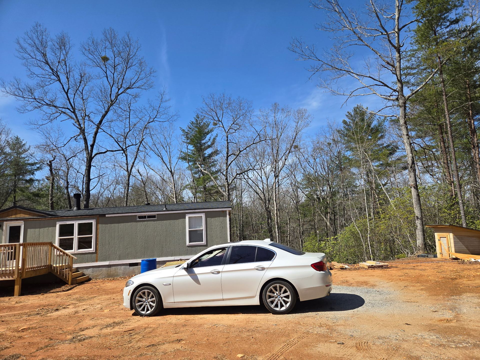 White car parked in front of a gray house in a wooded area on a sunny day.