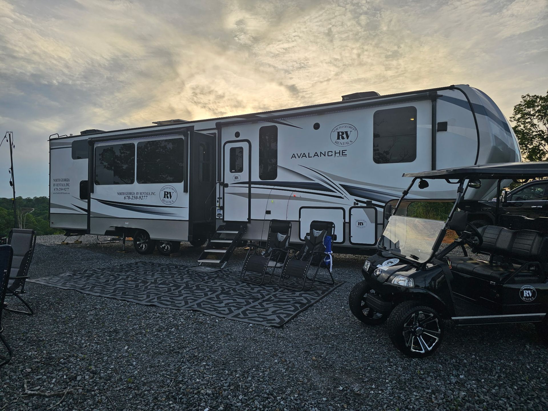 Large RV with slide-outs parked on gravel, golf cart beside it, under cloudy sky.