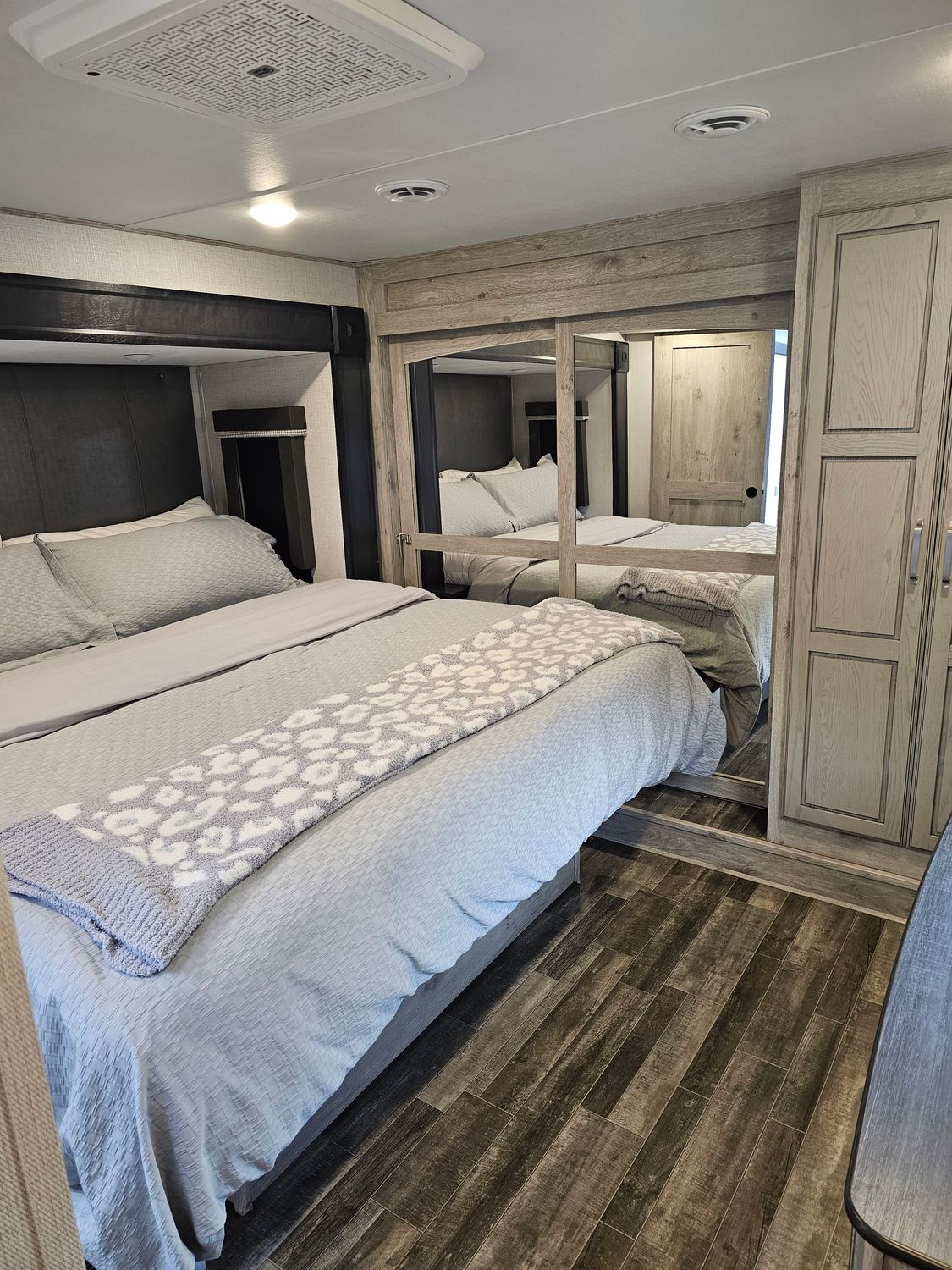 Bedroom in RV with bed, mirrored closet, and wood-look floor. Gray, white, and brown tones.