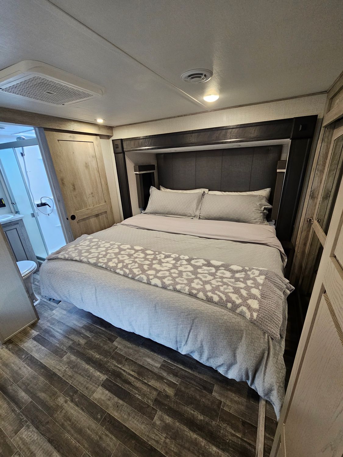 Bedroom in an RV with a queen bed, gray bedding, and a bathroom visible in the doorway