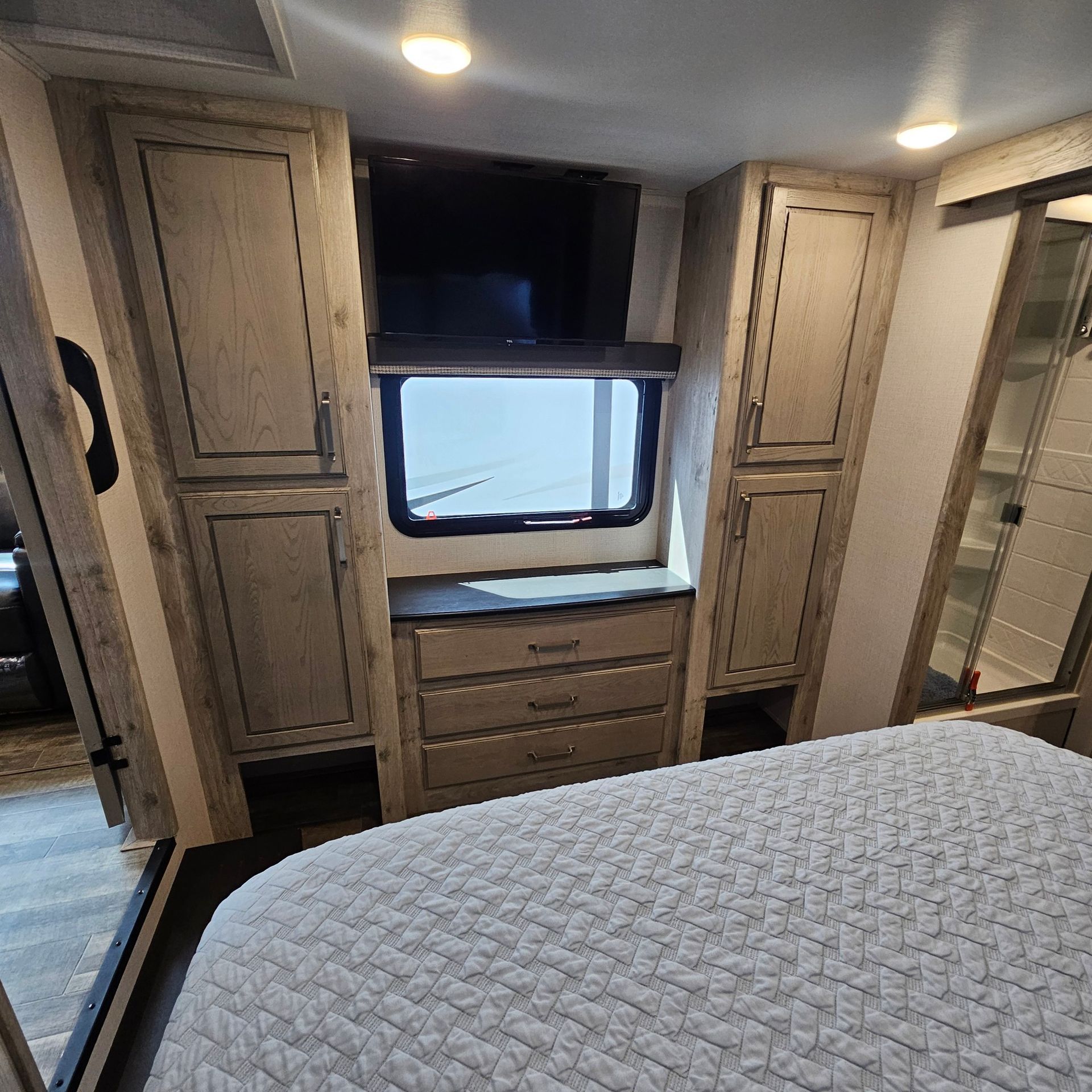 A bedroom in a RV with a bed, cabinets, drawers and a window
