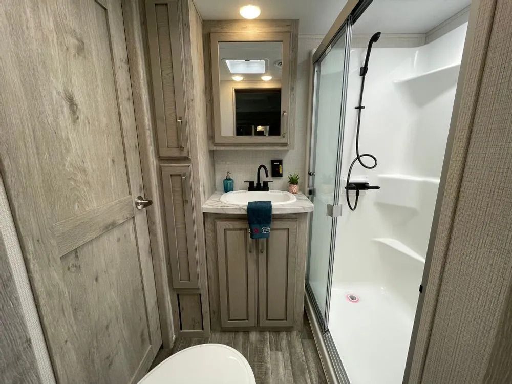 Bathroom in an RV: toilet, sink with cabinet, shower, tall storage, light wood-look walls