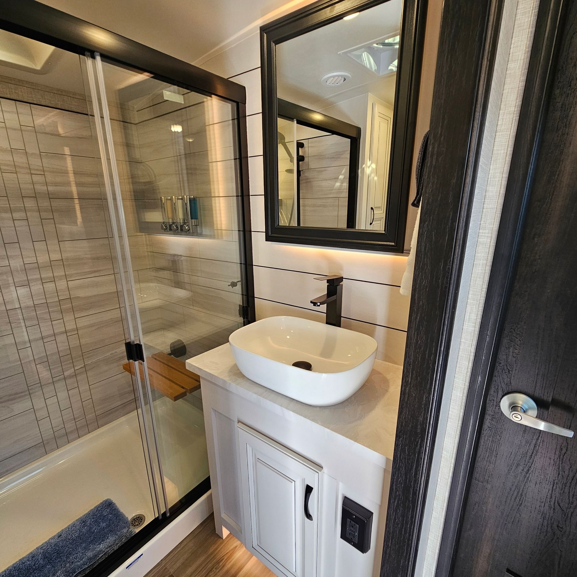 Modern RV bathroom with white vessel sink, black-framed mirror, glass shower, and light wood accents