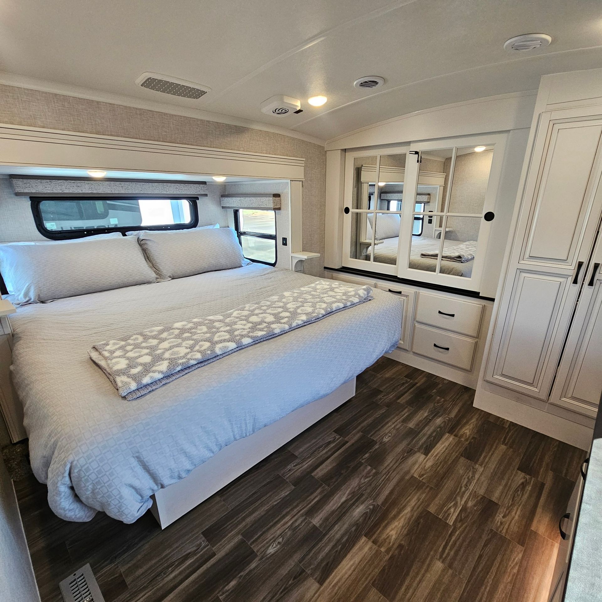 Bedroom interior of a recreational vehicle with a large bed, wardrobe, and dark wood-look flooring