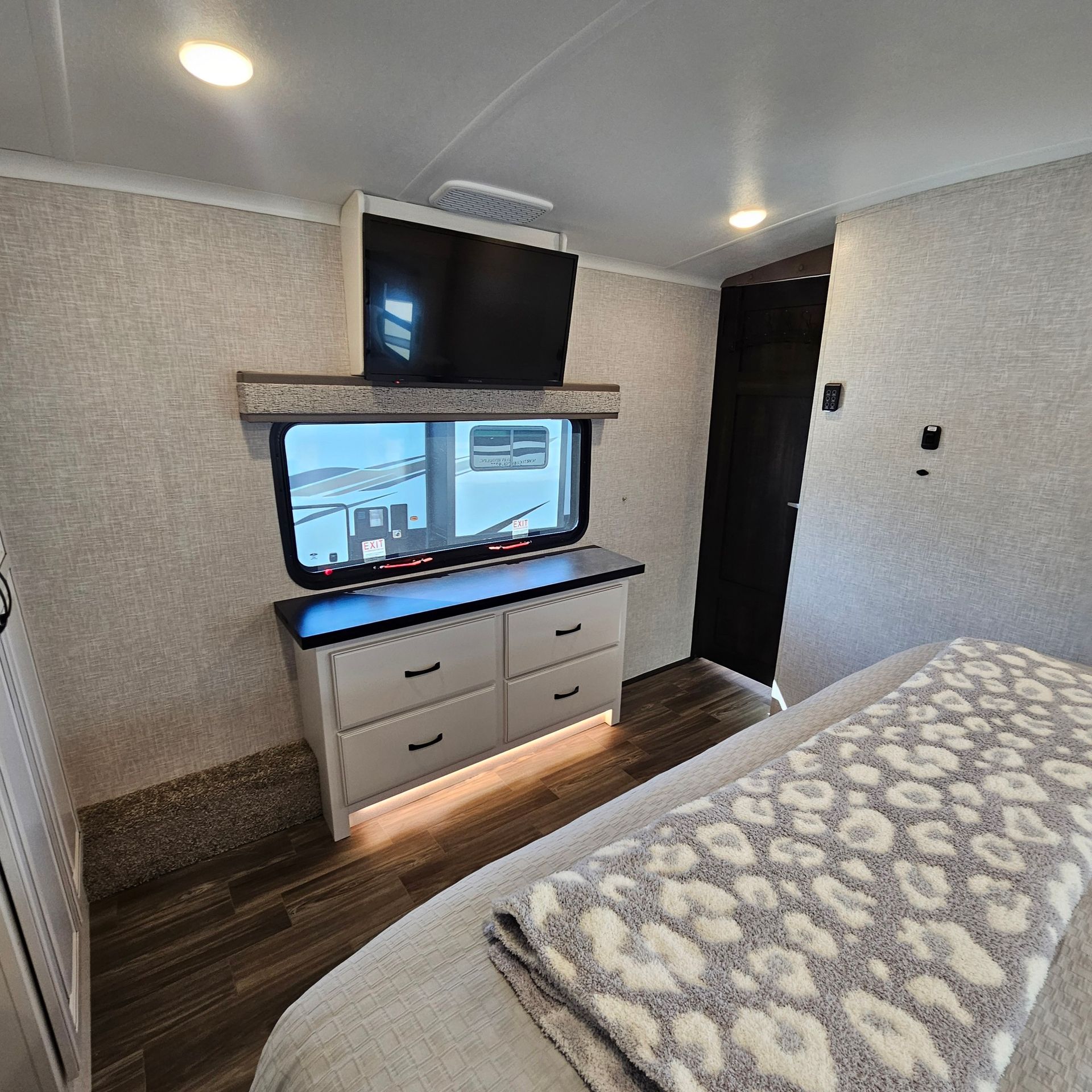Bedroom in a RV, with TV above a dresser, next to a door and bed with a throw blanket