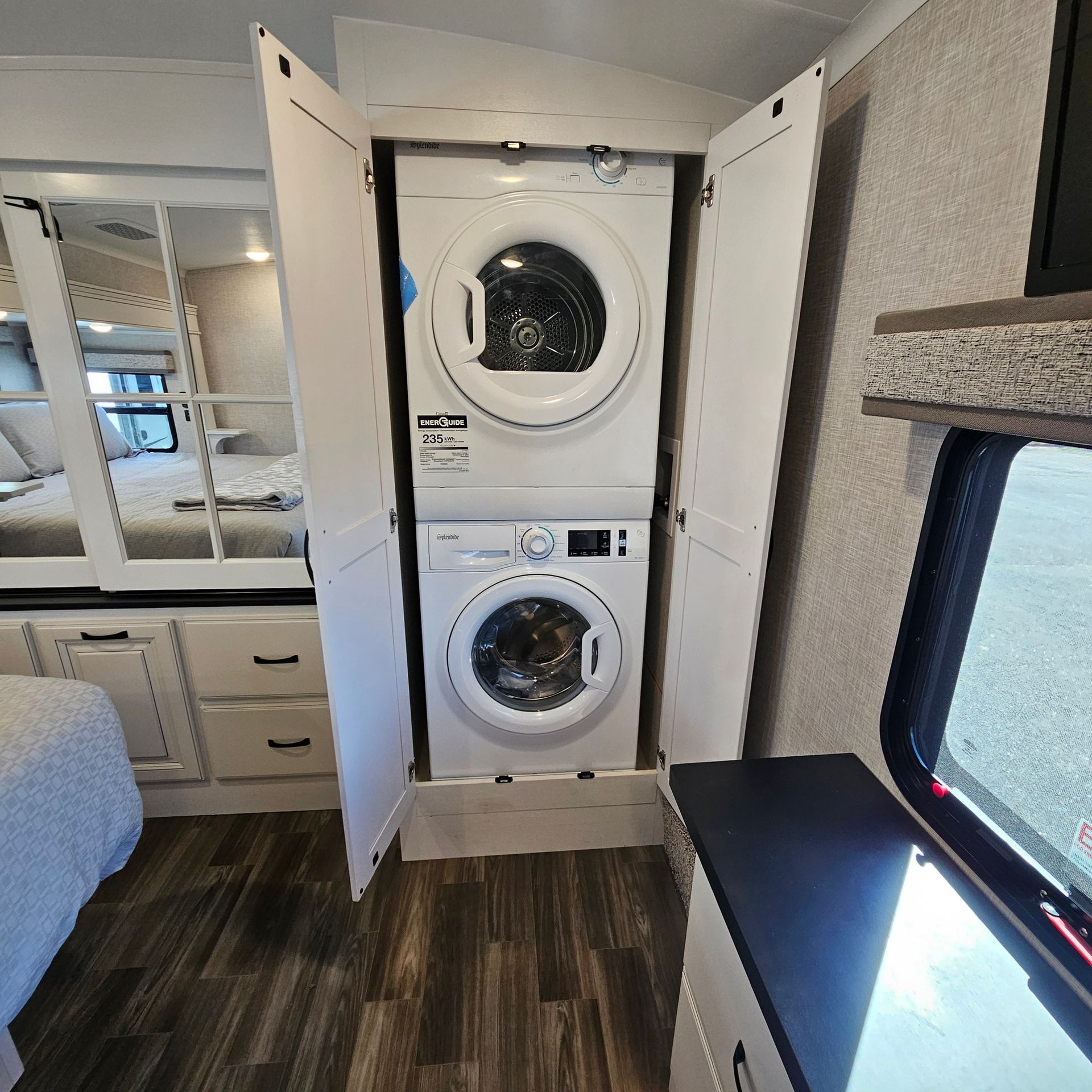 Stacked washer and dryer inside an RV, flanked by white cabinets, near a bed and window