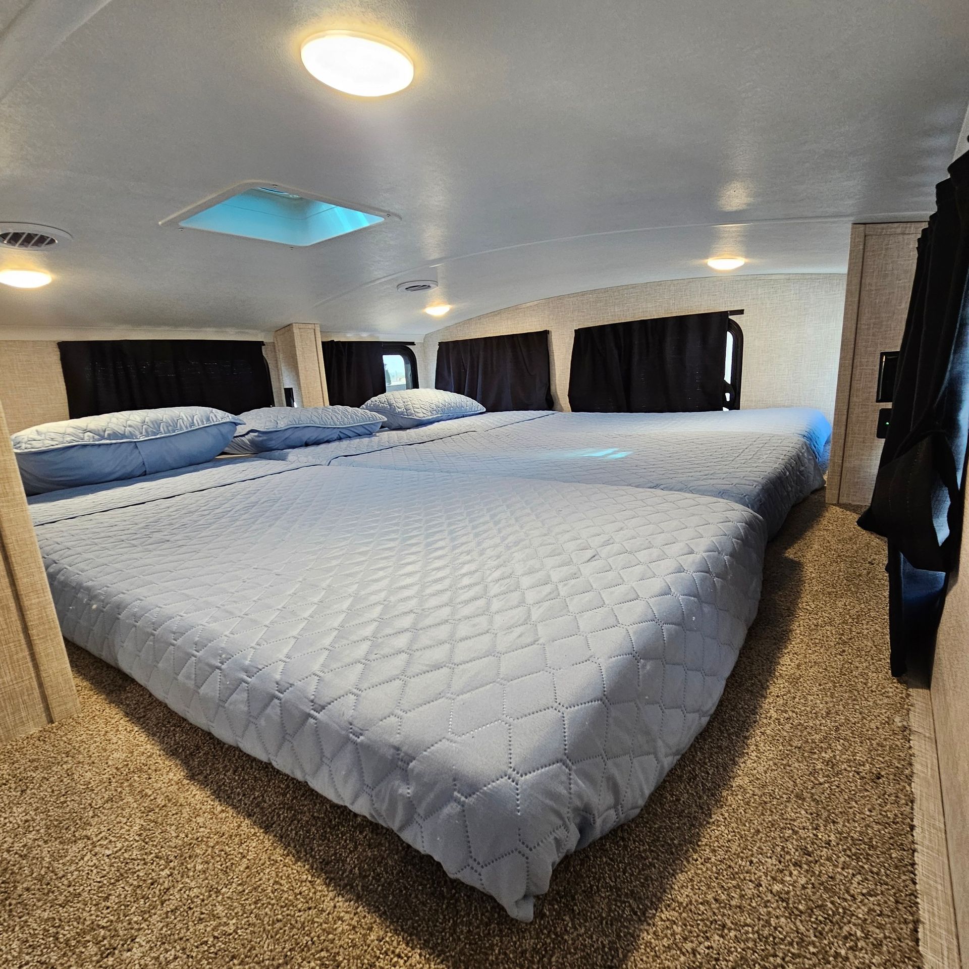Two large beds with blue bedding in a room, black curtains, and a skylight in the ceiling