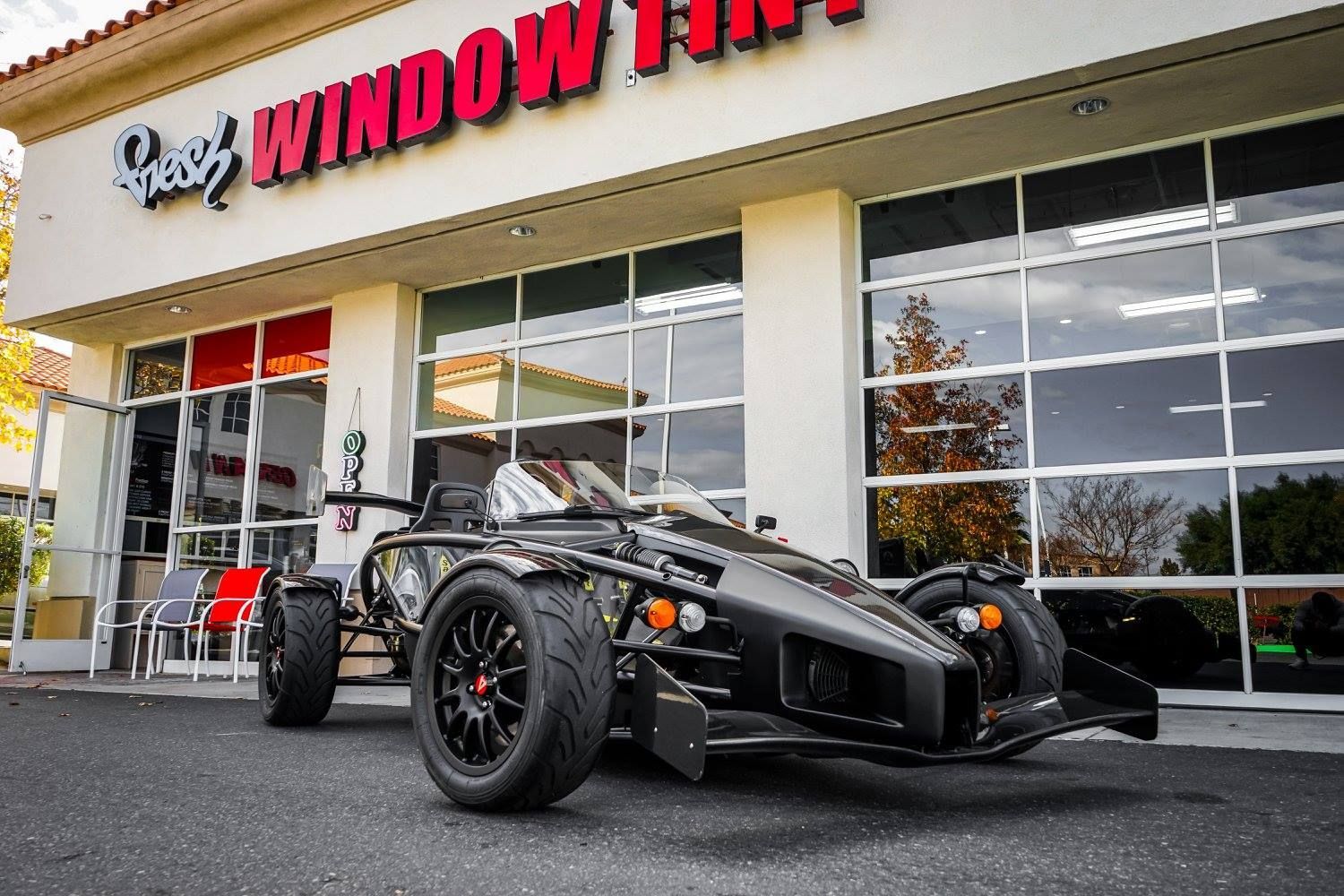 Black Ariel Atom sports car in front of a window tinting shop with reflective glass.