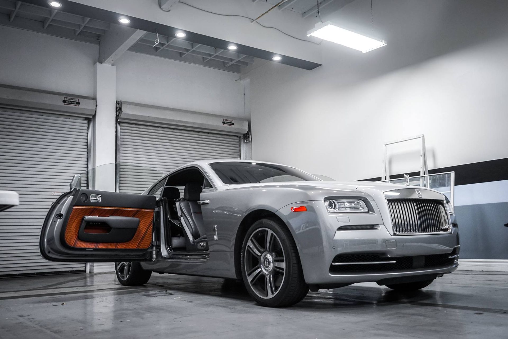 Silver Rolls-Royce coupe with open door in a garage setting with white walls and a roll-up door.