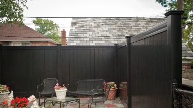 Backyard patio with black fence, wicker chairs, potted flowers, and a small table under a cloudy sky
