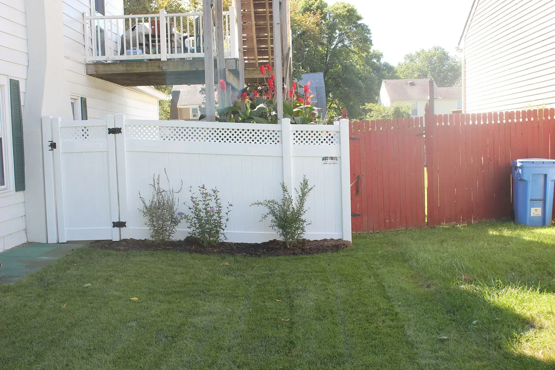 Backyard lawn with white fence, red gate, deck, shrubs, and blue recycling bin