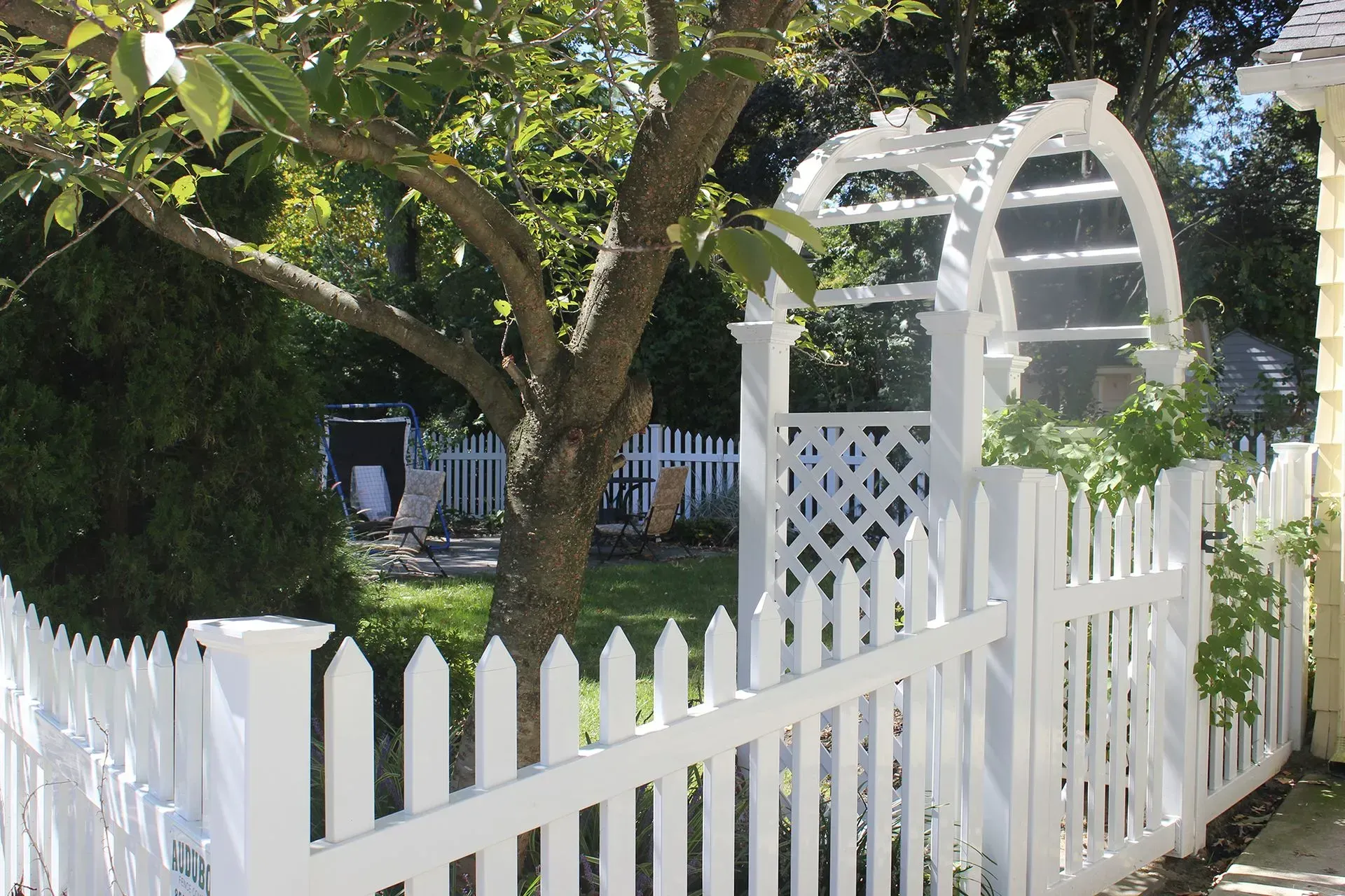 White picket fence and arbor around a leafy garden under trees in bright sunlight