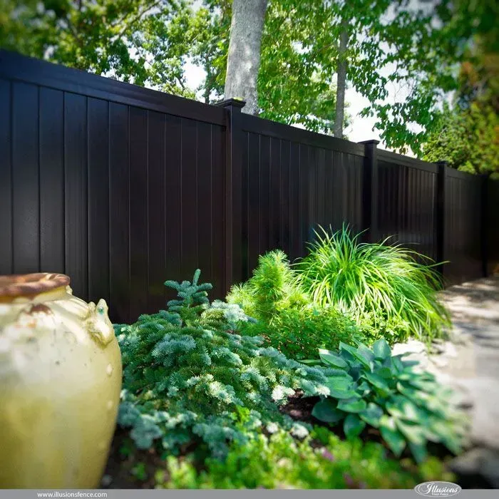 Backyard garden with dark privacy fence, lush green plants, and a large ceramic pot in sunlight