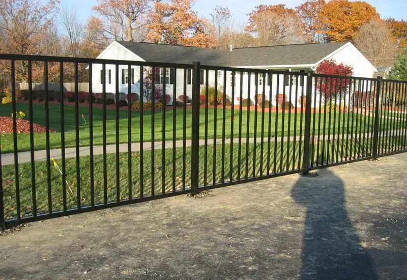 Black metal fence beside a driveway, with a white house and autumn trees in the background.