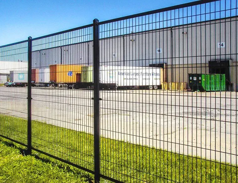 Black metal fence in front of industrial buildings, including a yellow shed and green dumpster.