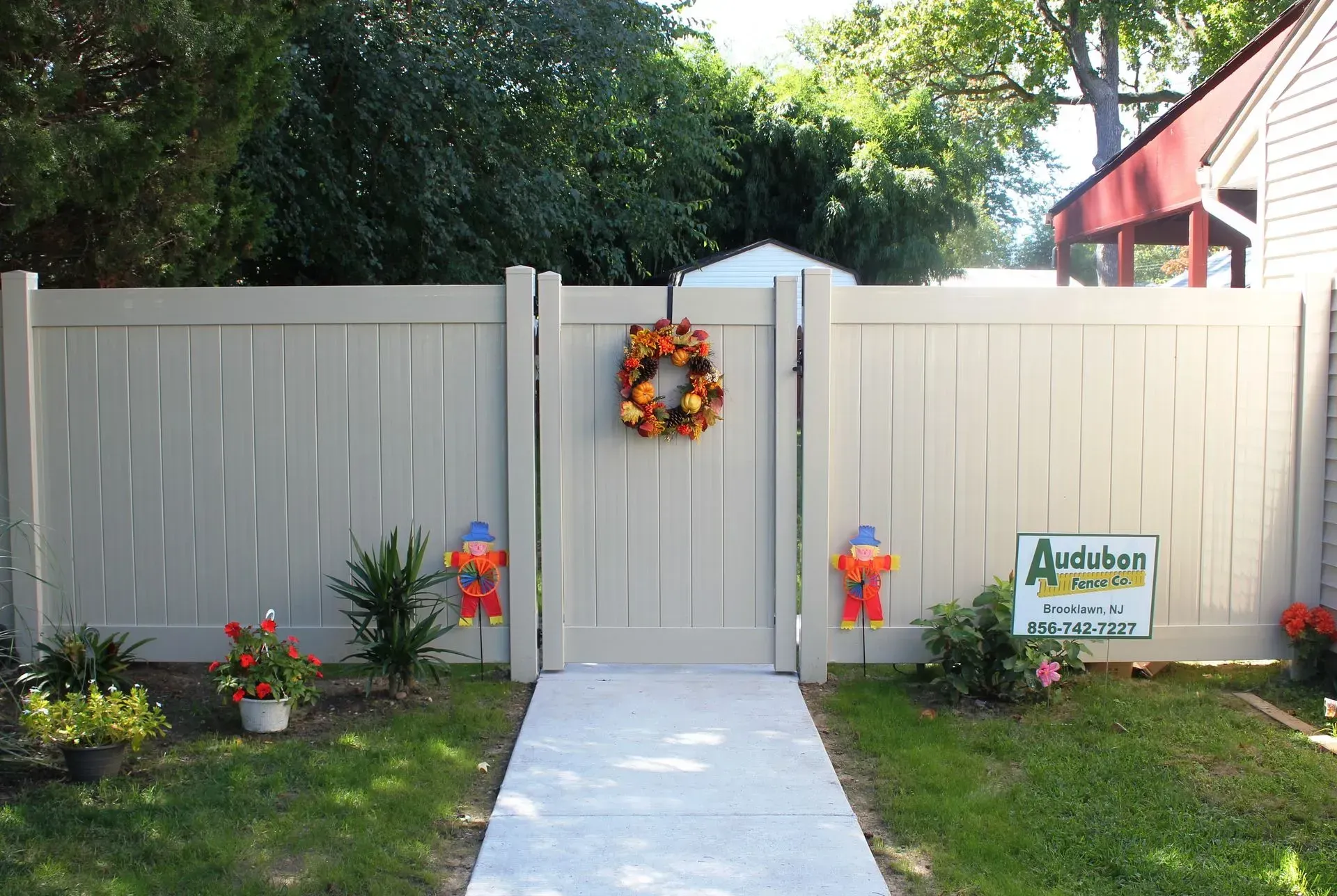Front gate with white fence, autumn wreath, and flower pots along a walkway in a sunny yard