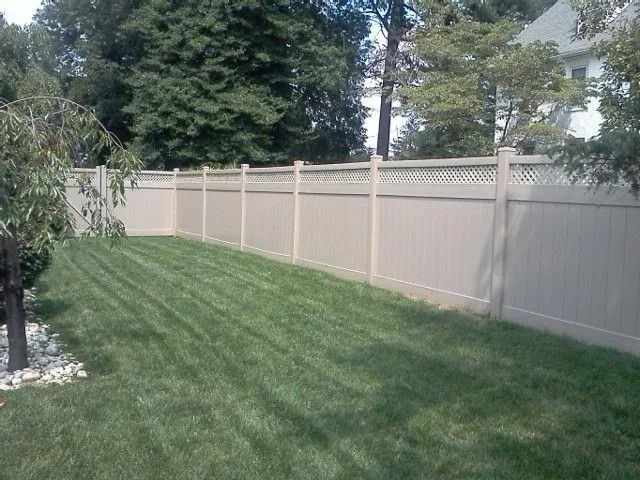 Backyard lawn beside a beige vinyl privacy fence with tall trees in the background