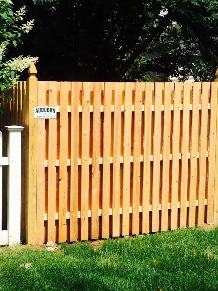 Wooden slat fence with a small sign, bordered by white posts and green grass in a sunny yard